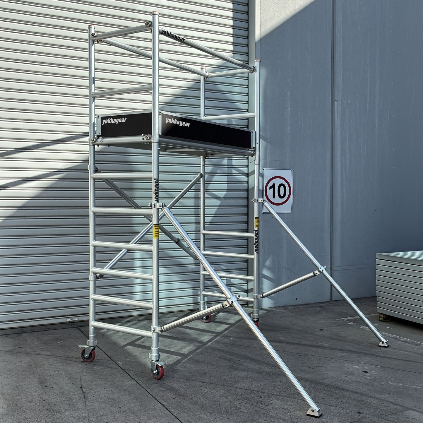 Yakka Gear Australia aluminium mobile scaffold shown in a warehouse, featuring 4m reachability, 2m platform height, 0.7m wide x 1.7m long platform, guard rails, outriggers and lockable wheels.