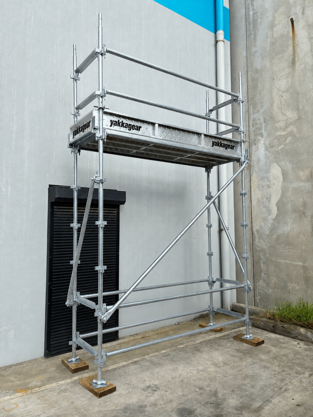Yakka Gear 5m reach slim Kwikstage Scaffolding tower in galvanised steel, featuring a 3m platform height and 0.7m slim width, with Yakka Gear branded platform components, safety guardrails, vertical standards, diagonal bracing and adjustable base jacks, shown assembled beside an industrial building for construction and elevated access applications across Australia.