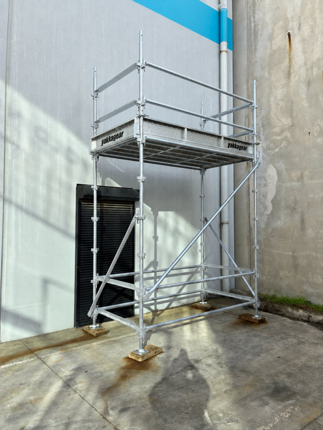 Yakka Gear 5m reach wide Kwikstage Scaffolding tower in galvanised steel, shown assembled on a concrete surface beside an industrial building wall in Australia, featuring a 3m platform height, safety guardrails, diagonal bracing, adjustable base jacks and Yakka Gear branded platform components, designed to provide stable elevated access for construction and maintenance work.