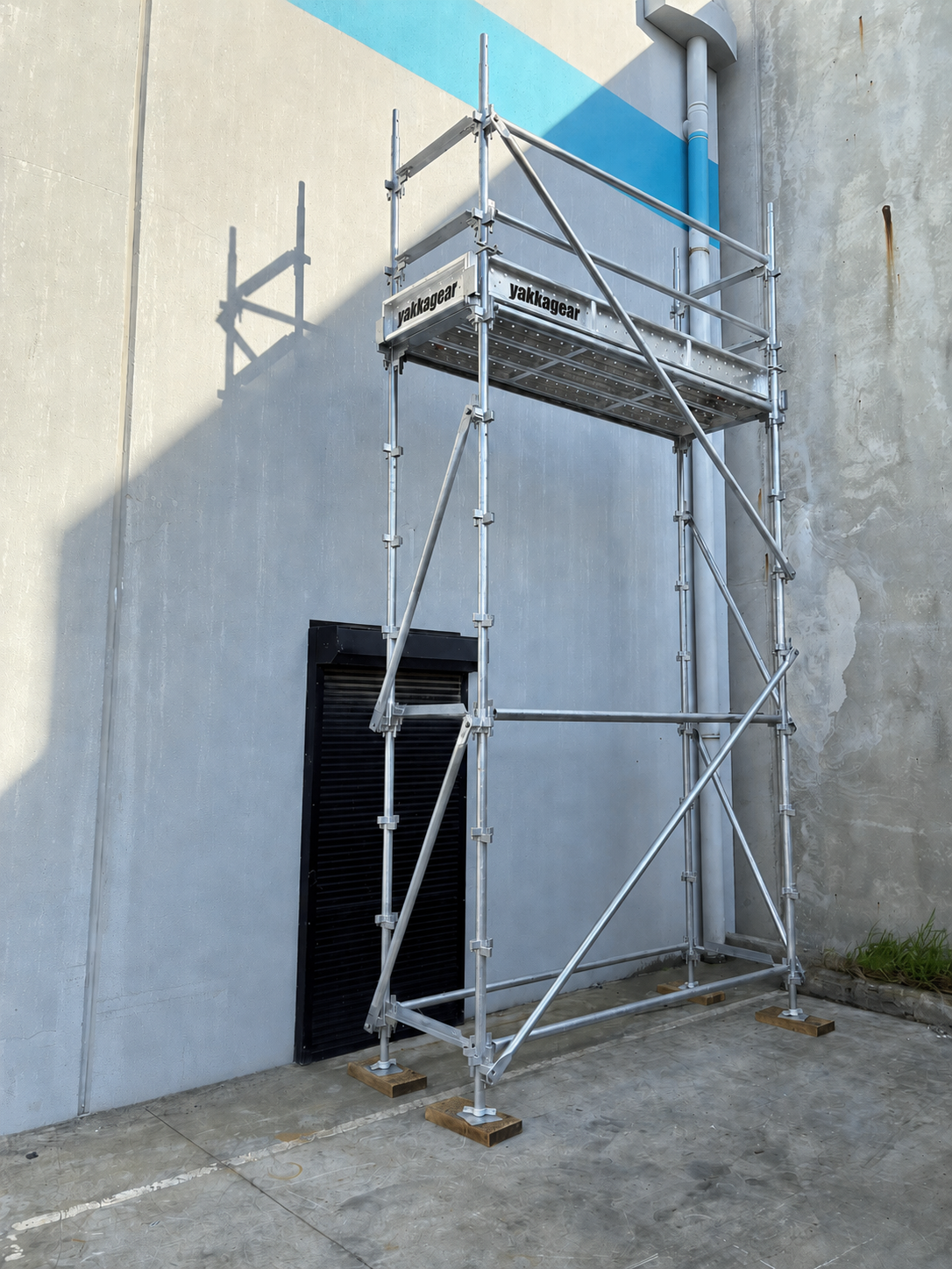 Yakka Gear 6m reach slim Kwikstage Scaffolding tower in galvanised steel, featuring a 4m platform height and 0.7m slim width, with Yakka Gear branded platform components, safety guardrails, vertical standards, diagonal bracing and adjustable base jacks, shown assembled beside an industrial building for construction and elevated access applications across Australia.