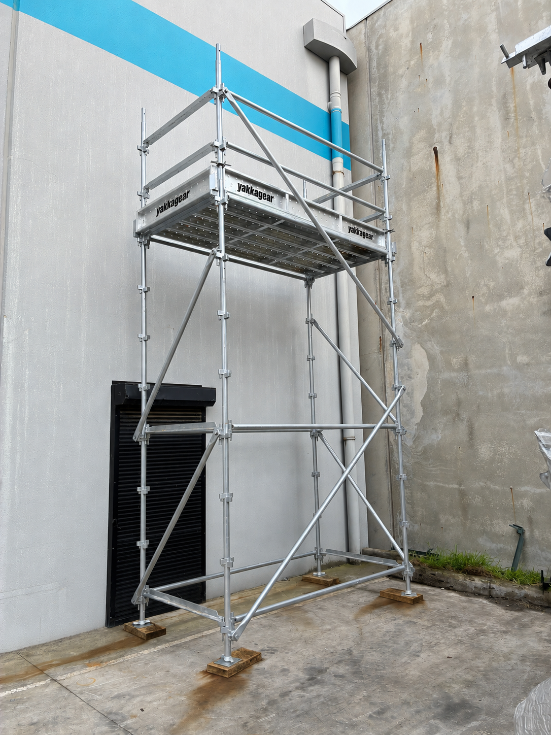 Yakka Gear 6m reach wide Kwikstage Scaffolding tower in galvanised steel, featuring a 4m platform height and 1.3m wide working platform, with Yakka Gear branded components, safety guardrails, vertical standards, diagonal bracing and adjustable base jacks, shown assembled beside an industrial building for construction and elevated access applications across Australia.