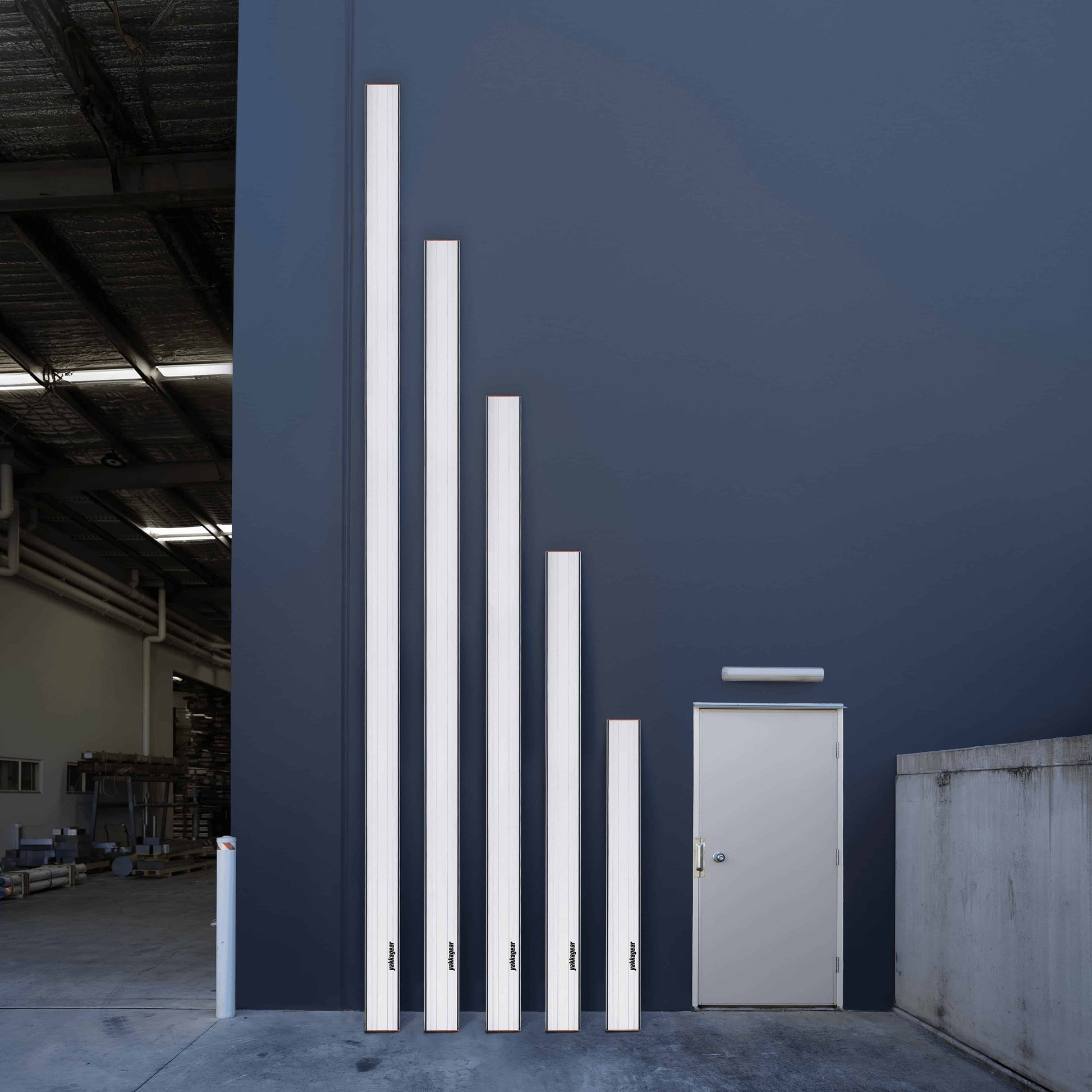 Yakka Gear aluminium planks in 2.0m, 3.0m, 4.0m, 5.0m, and 6.0m lengths lined up vertically against a warehouse wall, clearly displaying the size variations. All planks are made from 6063-T6 alloy with anti-slip surface, removable end caps, and a uniform profile of 228mm width x 45mm height x 2mm thickness.