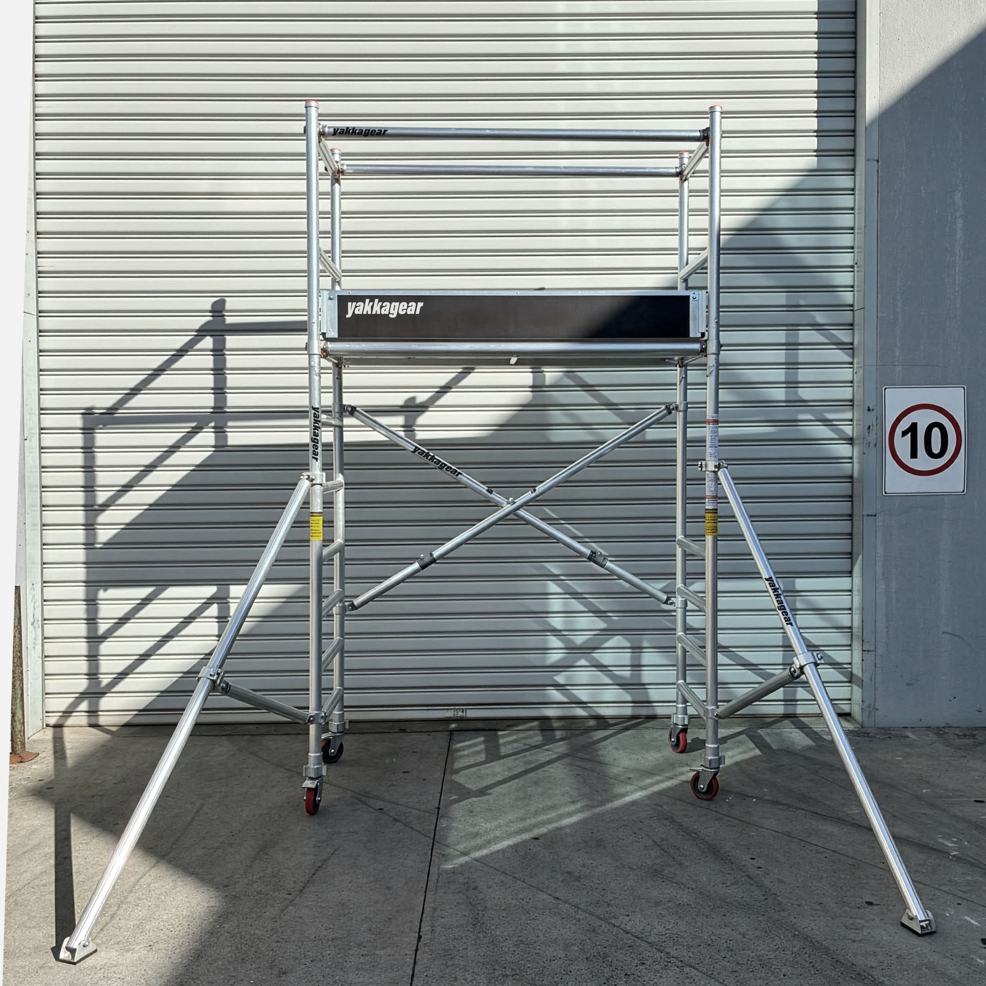 Yakka Gear Australia 4m reach aluminium scaffold tower in a warehouse setting, with a 2m platform height, 0.7m wide x 1.7m long deck, stabilising outriggers, guard rails and portable lockable wheels.