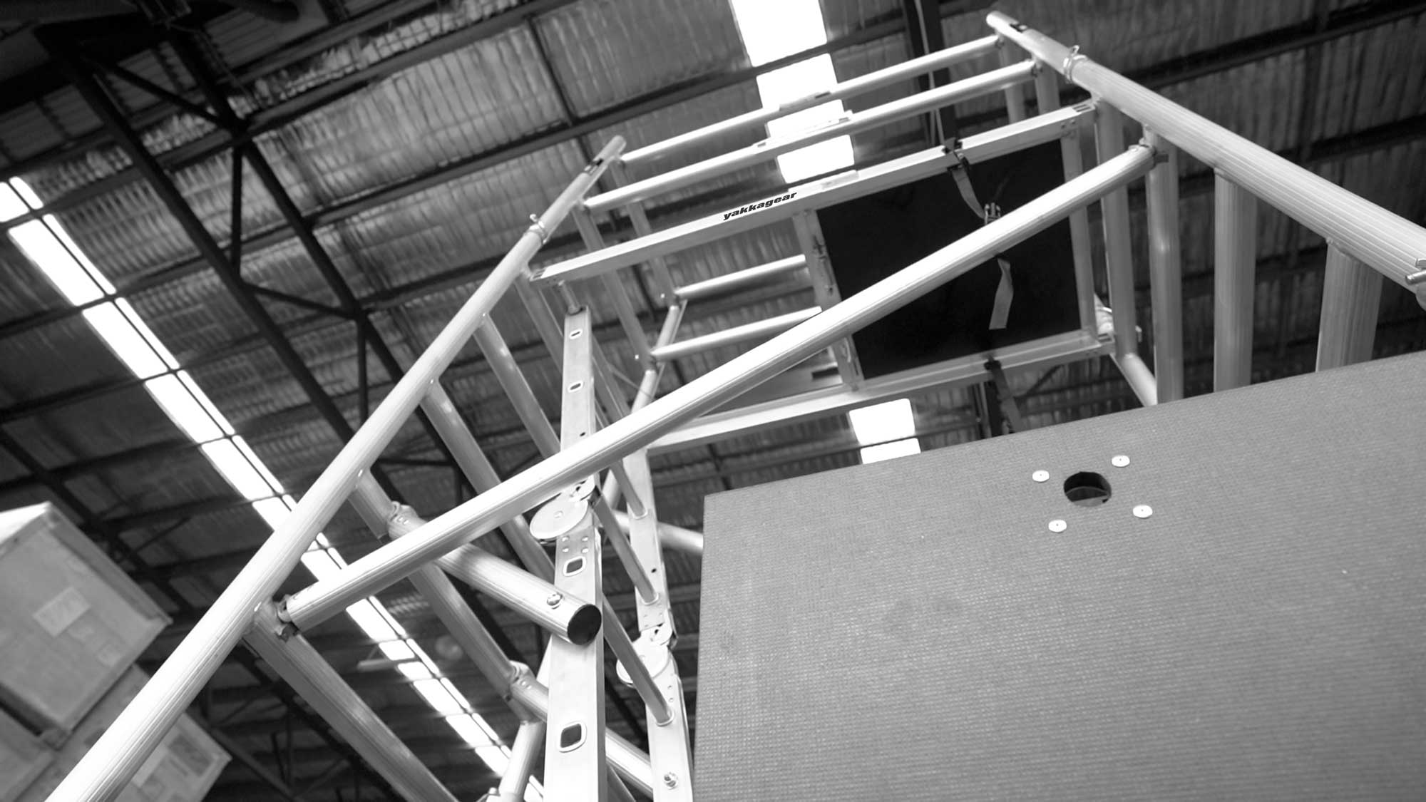 Yakka Gear Australia compact scaffolding tower as access equipment, including the Base Unit, Extension Unit, Tower Unit, and Adjustable Wheels. 1.44m Length x 0.74m Width, with working platform height of 3.9m and reach up to 6m high. View from ground level looking up towards the scaffolding set up in the warehouse. 